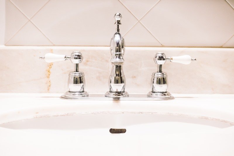 Close-up of a chrome bathroom faucet and handles installed on a white countertop, showcasing KOZ