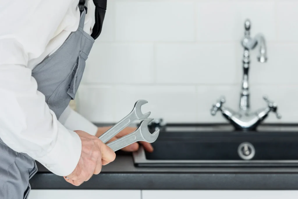 Plumber holding adjustable wrenches while working on a kitchen faucet, representing KOZ Plumbing & Heating's
