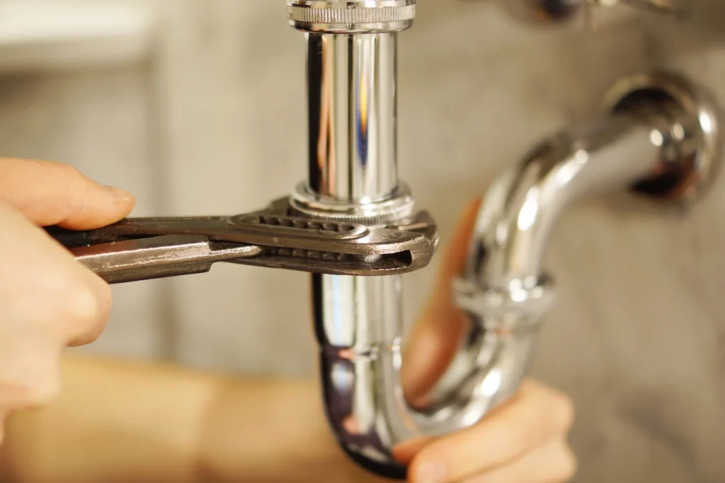 A plumber's hand uses pliers to repair a chrome pipe fitting, demonstrating emergency plumbing services